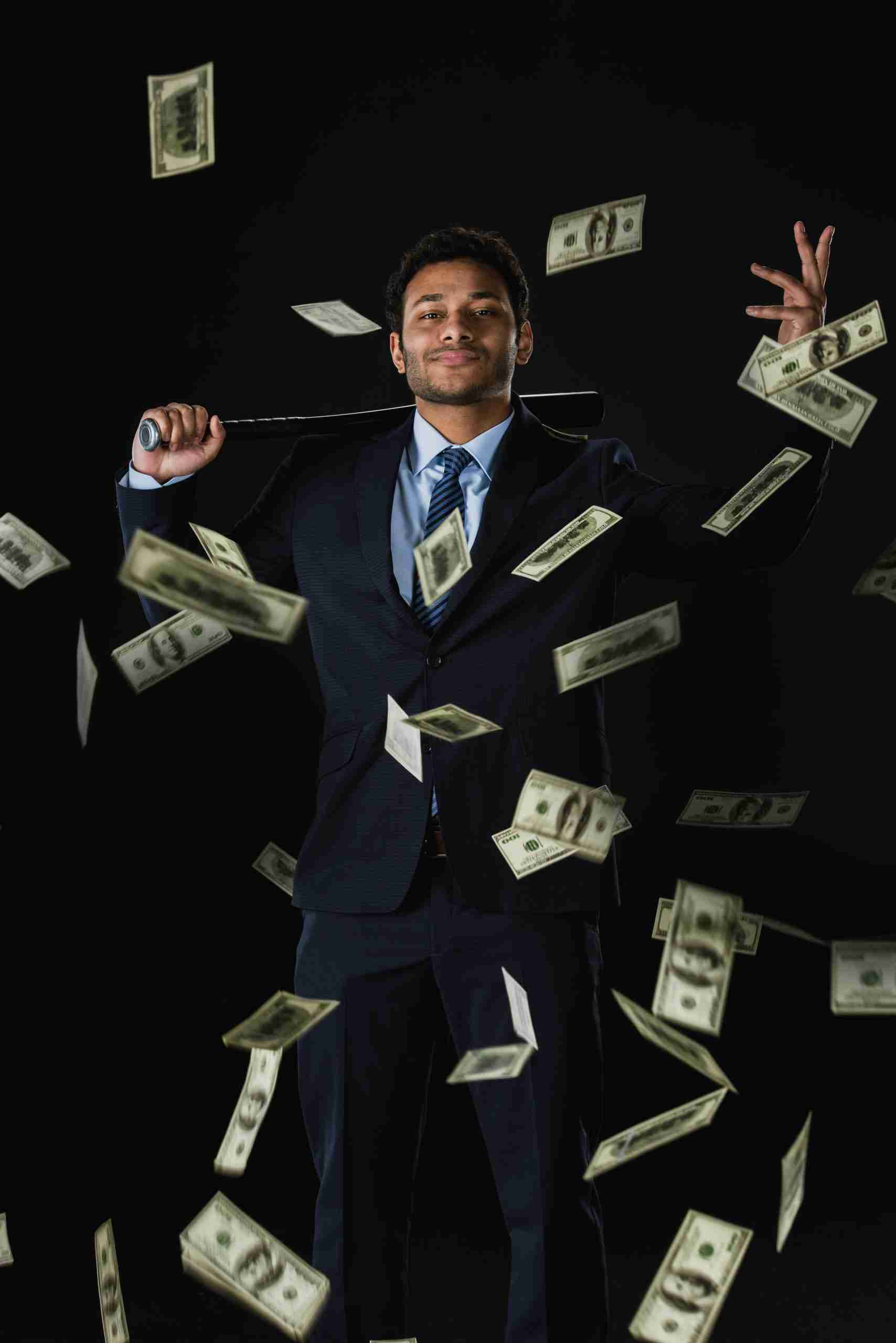 Confident young businessman holding baseball bat and dollar banknotes falling on black